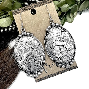 May include: Silver-toned oval earrings with a detailed relief of a horse in a mountainous landscape. The earrings have a decorative border and a simple hook closure. The design is reminiscent of Western or Southwestern jewelry.