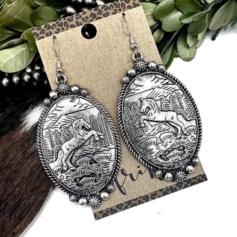 May include: Silver-toned oval earrings with a detailed relief of a horse in a mountainous landscape. The earrings have a decorative border and a simple hook closure. The design is reminiscent of Western or Southwestern jewelry.