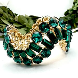 May include: A gold-toned cuff bracelet adorned with emerald green rectangular and faceted gemstones. The bracelet has a flexible design and is set against a white background with greenery.