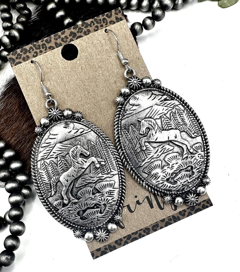 May include: Silver-toned oval earrings with a detailed relief of a rearing horse in a landscape. The earrings have a textured border and decorative accents. The design is set against a neutral background with a leopard print accent.