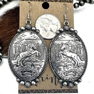 May include: Silver-toned oval earrings with a detailed western design. Each earring features a raised image of a horse rearing up in front of a forest and mountains. The earrings have a decorative rope-like border and floral accents.