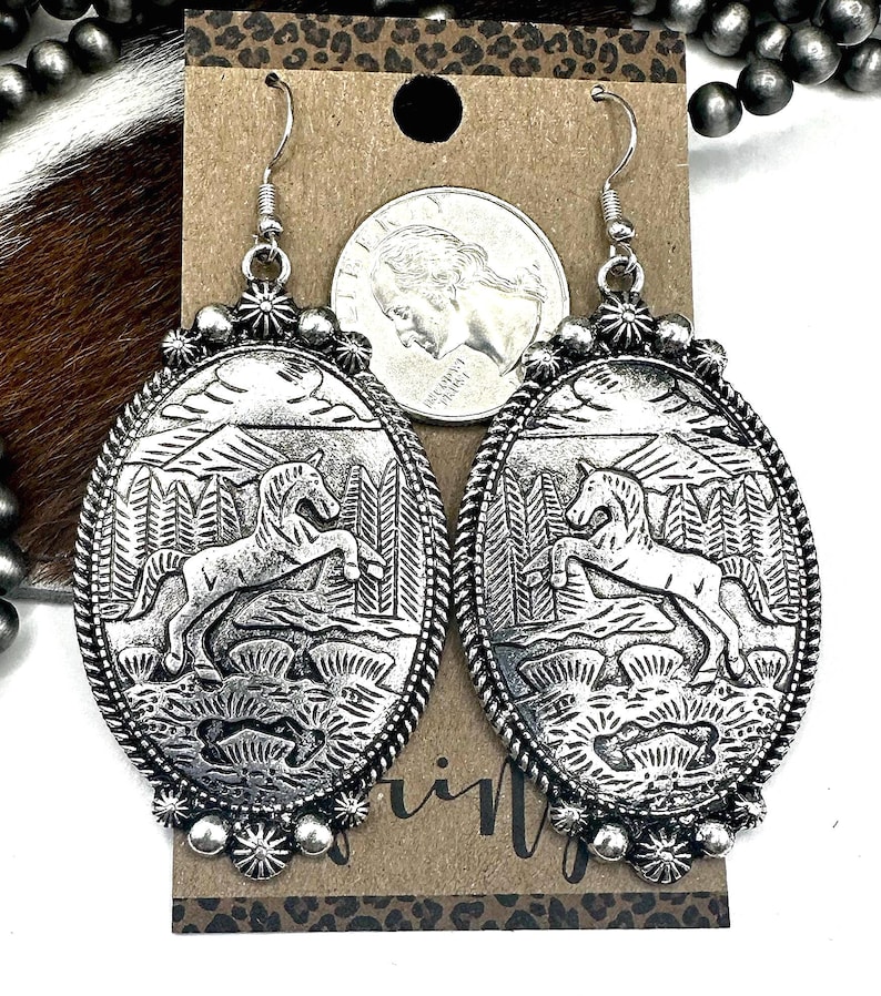 May include: Silver-toned oval earrings with a detailed western design. Each earring features a raised image of a horse rearing up in front of a forest and mountains. The earrings have a decorative rope-like border and floral accents.