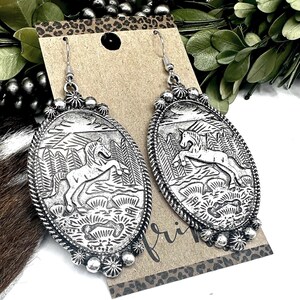 May include: Silver-toned oval earrings with a detailed relief of a horse in a landscape. The earrings have a decorative border and a floral design at the bottom. The design includes trees and a mountainous background.