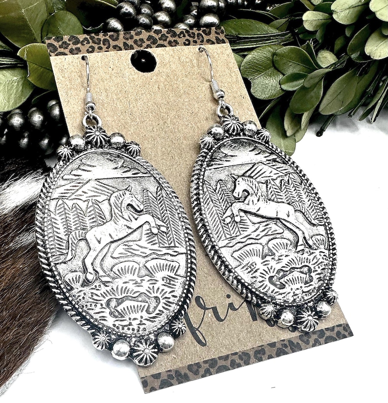 May include: Silver-toned oval earrings with a detailed relief of a horse in a landscape. The earrings have a decorative border and a floral design at the bottom. The design includes trees and a mountainous background.