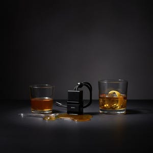 May include: A black square car air freshener with a black cord and the word HONEY printed on the front, positioned between a small glass of amber liquid and a glass of amber liquid with ice and a lemon peel, with a pool of honey on the surface.