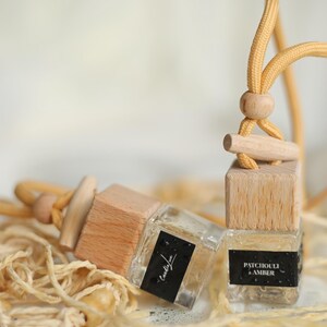 May include: Three small, clear glass bottles with wooden tops and yellow cords. Each bottle has a black label with white text. One label reads "Patchouli & Amber." The bottles are surrounded by beige twine.
