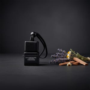 May include: A black car air freshener with the text "LAVENDER BLOOMS & SOOTHING SANDALWOOD". The freshener is suspended by a black cord. A small bundle of lavender, chamomile, and sandalwood sticks are next to the freshener.