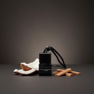 May include: A small square glass bottle with a black wooden cap and a black hanging cord. The label reads COCONUT SANDALWOOD & AMBER. The bottle is surrounded by pieces of raw coconut with brown husks and several small light brown wooden sticks.
