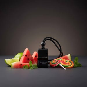 May include: A small square glass bottle with a black cap and a black hanging cord, labeled Watermelon & Lollipop. The bottle is surrounded by fresh watermelon slices, a lime wedge, green mint leaves, and two colorful swirl lollipops.
