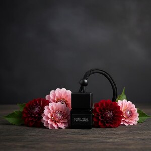 May include: A black square car air freshener bottle with a black cord and a label reading Dahlia Petals & Sandalwood. The bottle is surrounded by several dark red and light pink dahlia flowers with green leaves, arranged in a horizontal cluster.