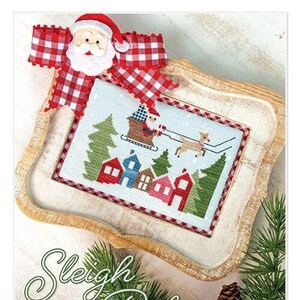 May include: A cross-stitch pattern depicting Santa Claus in a sleigh with reindeer flying over a winter village. The pattern is framed in a wooden frame with a red and white checked bow on top. The text "Sleigh Ride" is written below the frame. The website address "www.ItsSewEmma.com" is written below the text.