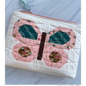 Puede incluir: Un estuche de mariposa acolchado con un diseño de mariposa rosa y verde azulado. El estuche es blanco con un patrón acolchado y cierre de cremallera. Tamaño final: 20 cm x 28 cm. El texto "Butterfly Pouch" está en la parte superior.