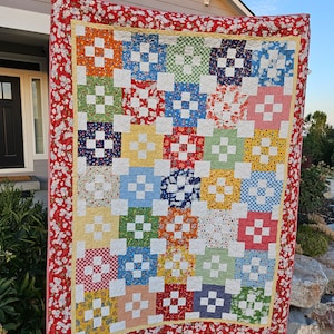 May include: A colorful handmade quilt with a patchwork design. The quilt features squares in various colors, including red, orange, yellow, blue, and green, with white cross-shaped accents. The border is red with a white floral pattern.