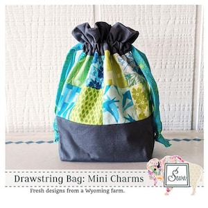 May include: A drawstring bag made from colorful patchwork fabric in shades of blue, green, and yellow. The bag has a gray bottom and is tied with blue and green cords. The text "Drawstring Bag: Mini Charms" and "Fresh designs from a Wyoming farm." is printed below the bag.