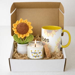 May include: A gift box with a sunflower theme. It contains a crocheted sunflower in a white pot, a candle with the text "Sending you Sunshine", and a mug with sunflower graphics and the word "vibes".