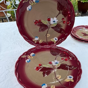 May include: A set of decorative ceramic plates with a floral design. The plates have a scalloped edge and a two-tone color scheme, with a burgundy rim and a tan center. The floral design features white flowers, red berries, and burgundy leaves.