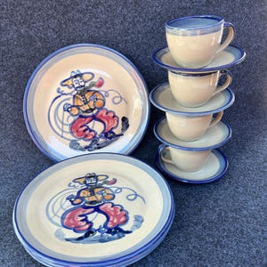 May include: A set of hand-painted ceramic dishes featuring a whimsical design. The set includes plates, saucers, and teacups. Each piece has a cream-colored base with a blue rim and a colorful illustration of a person. The teacups and saucers are stacked.