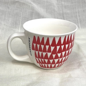May include: White ceramic coffee mug with a red geometric pattern. The mug has a large handle and the Starbucks logo on the side. The mug is sitting on a white surface.