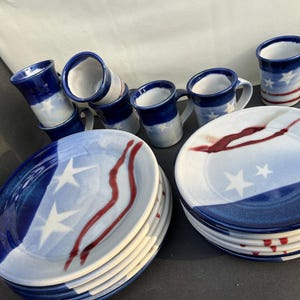 May include: A ceramic dinnerware set featuring a patriotic design. The set includes plates, mugs, and small cups. The plates and mugs have a blue and white color scheme with red accents and star details. The cups are blue and white.