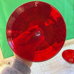 May include: A vibrant red glass bowl with a rounded shape. The bowl has a stem and a wide, shallow dish. The glass appears to have small bubbles throughout, giving it a textured appearance. The bowl is held up by a person.
