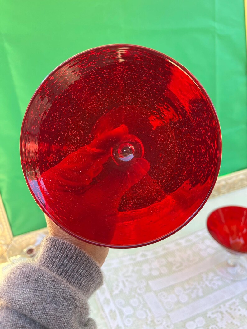 May include: A vibrant red glass bowl with a rounded shape. The bowl has a stem and a wide, shallow dish. The glass appears to have small bubbles throughout, giving it a textured appearance. The bowl is held up by a person.