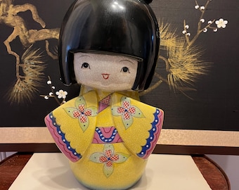 Könnte beinhalten: Eine japanische Kokeshi-Puppe aus Keramik mit glänzendem schwarzem Haar und einer roten Schleife. Die Puppe trägt einen gelben Kimono mit Blumenmustern und blau-rosa Besatz. Die Puppe steht auf einer weißen Oberfläche.