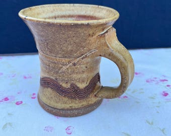 May include: A handmade ceramic mug with a tan and brown color scheme. The mug features a curved handle and a decorative wavy pattern around the middle. The mug is sitting on a floral patterned surface.