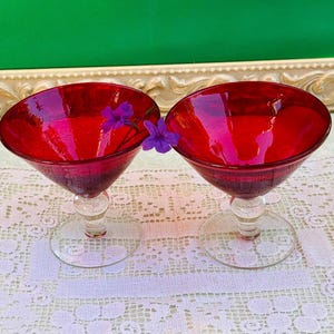 May include: Two ruby red cocktail glasses with clear stems and bases. The glasses are filled with light and are decorated with purple flowers. The glasses are on a white lace tablecloth.