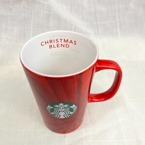 May include: Red Starbucks Christmas Blend 2014 mug with a white interior. The mug features the Starbucks logo and the words "CHRISTMAS BLEND" in red. The mug has a red handle and a red exterior with a subtle pattern.