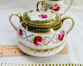 May include: A white porcelain sugar bowl and creamer set with a green and gold floral design. The set features pink roses and gold accents.