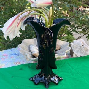 May include: A glossy black ceramic vase with a flared, scalloped rim and a textured, leaf-like vertical design on the body. The vase stands on a wide, multi-pointed base and contains several white flowers with delicate pink streaks on the petals.