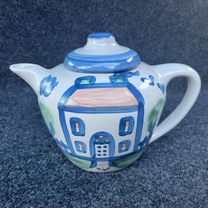 May include: A ceramic teapot with a white base and a blue lid. The body features a hand-painted design of a house with a peach-colored roof, blue outlines, and green foliage accents. The teapot includes a curved handle and a short, tapered spout.
