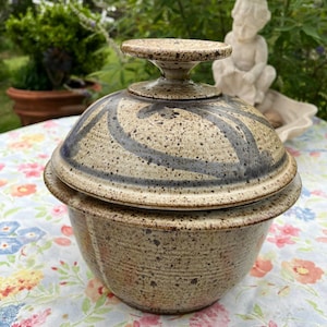 May include: A ceramic lidded bowl with a speckled beige and brown finish. The lid features a dark blue painted floral pattern and a flat, circular handle. The bowl has a wide rim and a rounded base, showcasing a textured, earthy appearance.