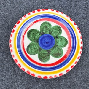 May include: A round, hand-painted ceramic plate with a floral design. The plate features a green flower in the center with a blue center, surrounded by red, blue, and yellow concentric rings. The rim has red dots.