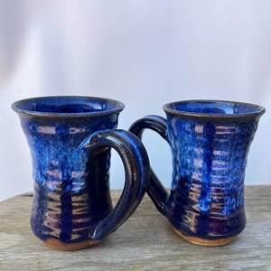 May include: Two handmade ceramic mugs with a deep blue glaze. The mugs have a textured surface and curved handles. The interior and exterior are a rich, glossy blue, with a darker blue at the base. The mugs are on a wooden surface.
