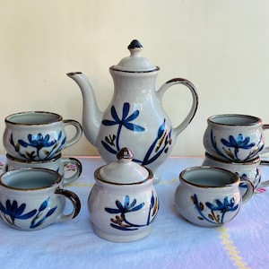 May include: A vintage ceramic tea set with a teapot, sugar bowl, and six cups. The set features a floral design with blue flowers and brown stems on a white background.