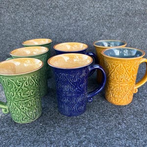 May include: A collection of ceramic mugs in various colors, including green, blue, and yellow. Each mug features a textured, embossed design and a contrasting interior. The mugs have curved handles and are suitable for hot beverages.