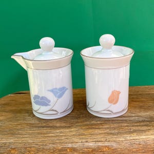 May include: Two white ceramic serving pieces with lids, one with a spout. Each piece has a beige band and a floral design in blue and orange. The pieces are on a wooden surface.
