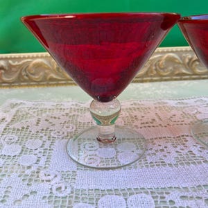 May include: Two ruby red martini glasses with a clear stem and base. The glasses are set on a white lace tablecloth. The background is a green surface.