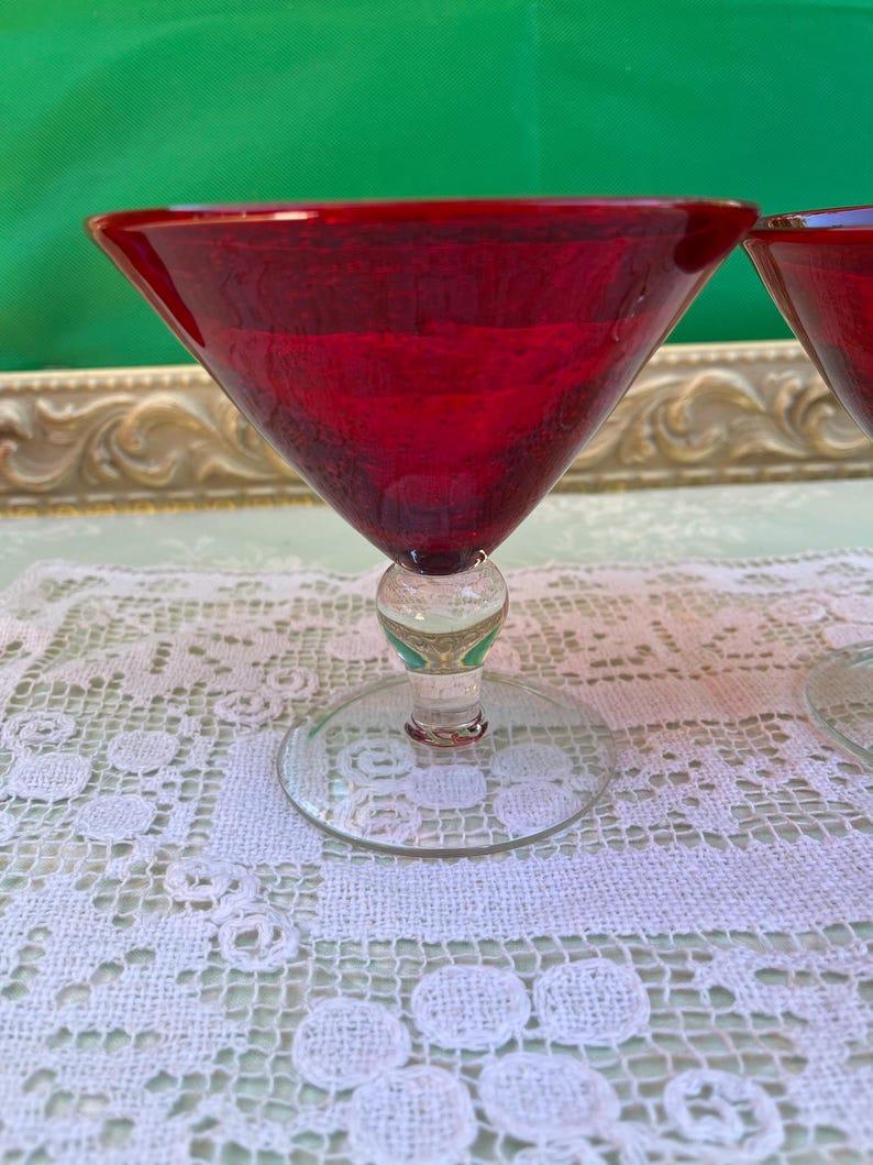 May include: Two ruby red martini glasses with a clear stem and base. The glasses are set on a white lace tablecloth. The background is a green surface.