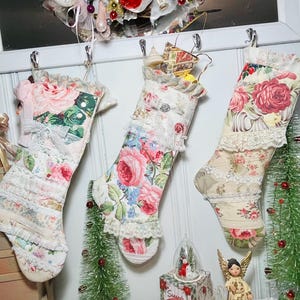 May include: Three white Christmas stockings with floral patterns and lace trim. The stockings are hung on a white wall with hooks. The stockings are filled with small gifts and ornaments.