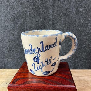 May include: A small, off-white ceramic mug with a blue rim and handle. The mug is inscribed with the words "Wonderland of Lights" in blue script, along with the year "1993". The mug sits on a small, polished, reddish-brown wooden block.