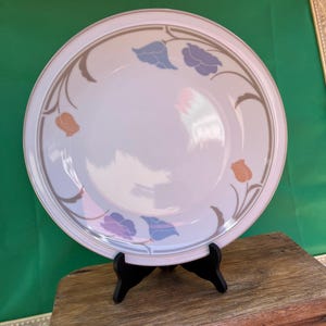 May include: White ceramic plate with a floral design featuring blue, purple, and orange tulip-like flowers. The plate has a brown border and is displayed on a black stand. A decorative piece for the dining table.