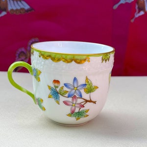 May include: A white porcelain teacup with a green rim and handle. The cup is decorated with a floral pattern in shades of pink, blue, green, and yellow.