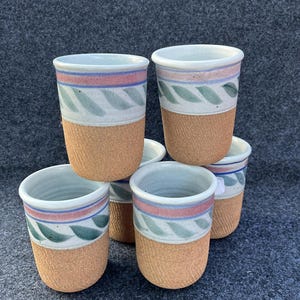 May include: A stack of six ceramic cups. The cups have a textured, light brown base and a white top with a blue, pink, and green leaf design. The interior of the cups is white.