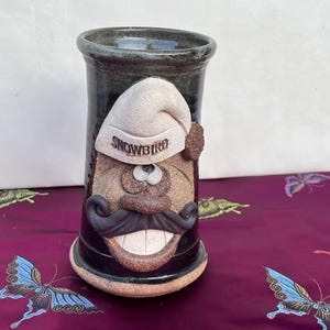 May include: A ceramic vase with a whimsical face, featuring a white sleeping cap with the word "SNOWBIRD" on it, a brown mustache, and a wide, toothy smile. The vase is glazed in shades of brown, green, and white.