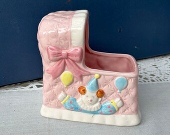 May include: A pink ceramic baby crib with a white trim and a pink bow. A clown with a blue hat and red hair is holding balloons. The crib is decorated with a white and pink quilted pattern.