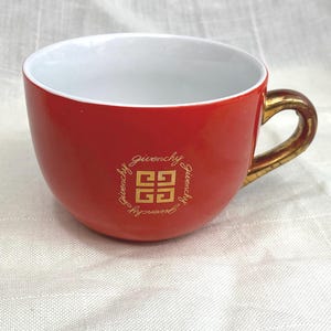 May include: Red ceramic teacup with a gold handle and the Givenchy logo in gold. The interior of the cup is white. The cup is sitting on a white surface.