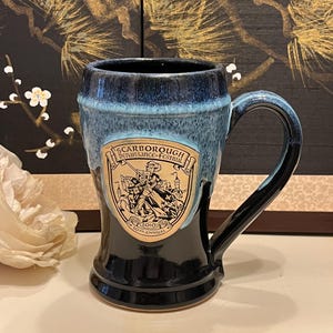 May include: A ceramic mug with a black base and a blue-glazed top. The mug features a handle and a shield-shaped emblem with the text "Scarborough Renaissance Festival" and "30th Annual".
