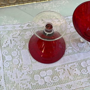 May include: Two vintage red glass goblets with clear stems and bases. The goblets are set on a white lace tablecloth. The goblets have a rounded bowl shape. The background includes a decorative gold-framed mirror.